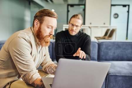 bearded entrepreneur showing laptop with startup project to strict businessman in black turtleneck pointing with finger while sitting in lounge of modern office, business partnership concept