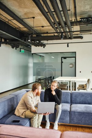 stylish business partners discussing startup project while sitting on comfortable couch with laptop in lounge of coworking office with high tech interior, business partnership concept