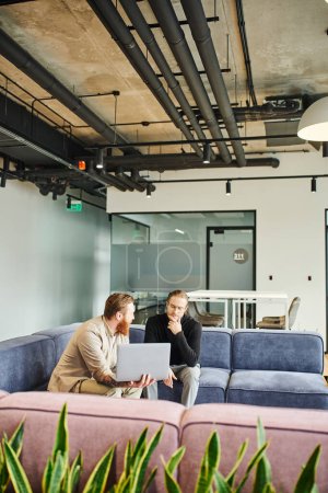 bearded businessman showing startup project on laptop to thoughtful colleague while talking on comfortable couch in lounge of modern office with high tech interior, business partnership concept