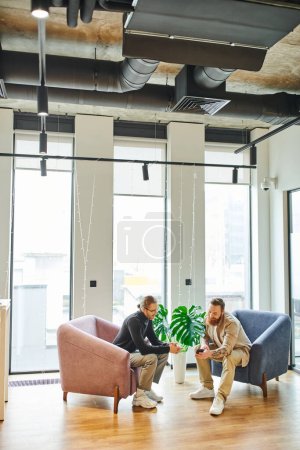 full length of bearded man showing smartphone to colleague in black turtleneck while sitting in office lounge with cozy armchairs and large windows, partnership and success concept