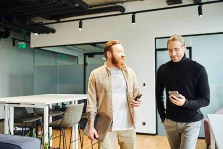 cheerful, bearded and tattooed businessman with laptop talking to smiling colleague looking at mobile phone while standing in modern office, productivity and cooperation concept