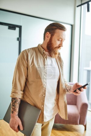 bearded and tattooed businessman in stylish casual clothes holding laptop and networking on mobile phone while standing in modern office, successful entrepreneurship concept