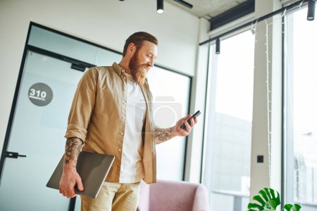 pleased, bearded and tattooed businessman in stylish casual clothes browsing internet on mobile phone while standing with laptop in modern coworking office, successful entrepreneurship concept