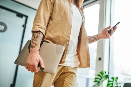 Photo for Partial view of tattooed entrepreneur in stylish casual clothes standing with laptop and messaging on mobile phone in modern office environment, successful business concept - Royalty Free Image