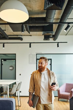 cheerful, tattooed and bearded businessman with laptop and mobile phone standing and looking away in contemporary office environment with high tech interior, successful entrepreneurship concept