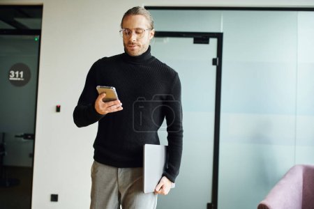 thoughtful and stylish businessman in eyeglasses and black turtleneck holding laptop and looking at mobile phone in modern office environment, successful business concept