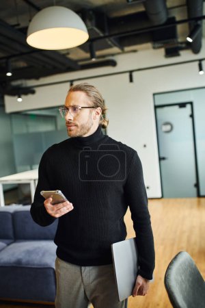 serious businessman in black turtleneck and stylish eyeglasses looking away while standing with laptop and smartphone in modern coworking environment, successful business concept