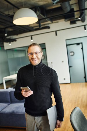 professional headshot of cheerful businessman in black turtleneck holding laptop and smartphone while looking at camera in modern office with high tech interior, successful business concept