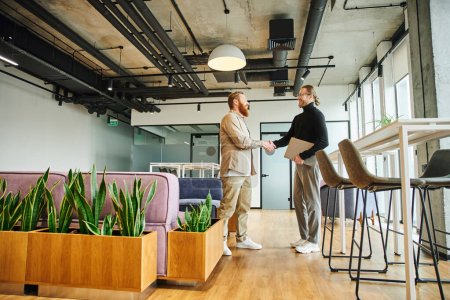Photo for Side view of stylish businessman holding laptop and shaking hands with tattooed and bearded colleague in lounge of coworking environment with modern furniture and floral decor - Royalty Free Image