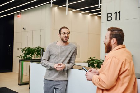 Aufgeregter Geschäftsmann in Brille und lässiger Kleidung im Gespräch mit bärtigen Kollegen am Empfang in der Lobby eines modernen Coworking-Büros, Unternehmer diskutieren über die Möglichkeit eines Start-ups