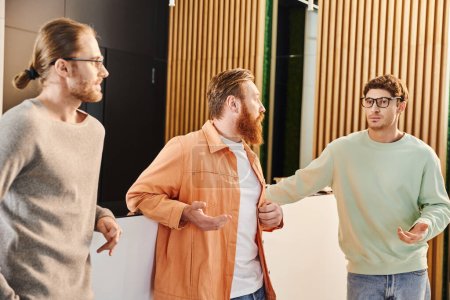 bearded businessman talking to serious colleagues in eyeglasses and casual clothes while planning startup project in lobby of contemporary coworking environment, business collaboration