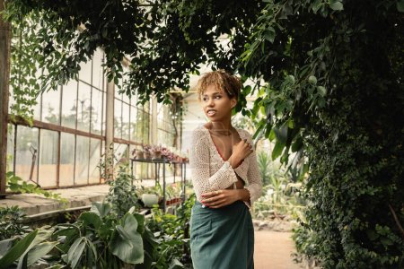 Mujer joven afroamericana sonriente y de moda en verano tejida arriba y falda mirando hacia otro lado mientras está de pie cerca de plantas verdes en el centro del jardín, mujer elegante disfrutando de un exuberante entorno tropical