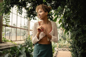 Positive young african american woman in trendy summer knitted top and skirt looking away while standing near green plants in garden center, stylish woman enjoying lush tropical surroundings Poster #663909254