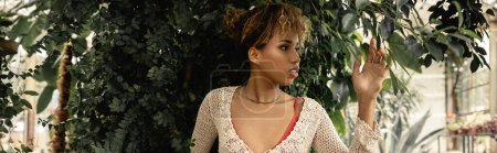 Photo for Young and trendy african american woman with makeup and knitted top touching green plants while standing in blurred garden center, stylish woman enjoying lush tropical surroundings, banner - Royalty Free Image