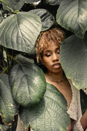 Mujer afroamericana joven de moda con maquillaje usando top de punto mientras está de pie cerca de hojas verdes borrosas en invernadero, mujer elegante disfrutando de un exuberante entorno tropical