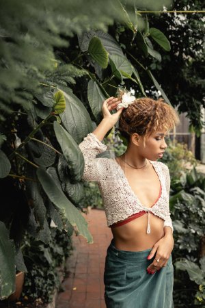 Modische junge afrikanisch-amerikanische Frau in Stricktop und Rock, die die Haare berührt und neben grünem Laub im verschwommenen Indoor-Garten steht, modebewusste Dame in tropischer Oase