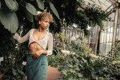 Confident young african amerian woman in knitted top and skirt looking away while standing near plants and leaves in blurred garden center, summer fashion-forward lady in tropical oasis Stickers #663909680