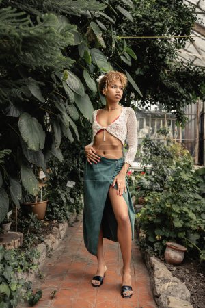 Full length of trendy young african american woman in skirt and knitted top looking away and standing near green plants in greenhouse, stylish lady surrounded by lush greenery, summer