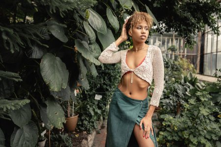 Foto de Mujer afroamericana joven de moda en traje de verano y la parte superior de punto posando cerca de plantas verdes y de pie en el jardín interior borroso, señora elegante rodeada de exuberante vegetación, verano - Imagen libre de derechos