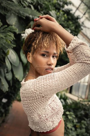 Portrait de jeune femme afro-américaine confiante en tricot haut et tenue d'été posant et debout près de plantes floues en orangerie, fashionista se mélangeant à la flore tropicale