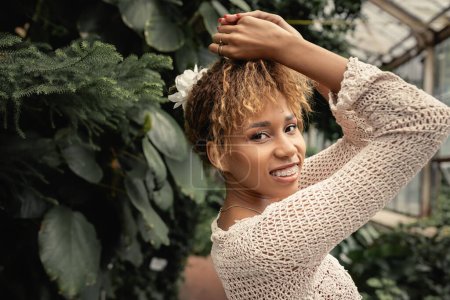 Portrait de jeune femme afro-américaine souriante avec bretelles et tenue d'été regardant la caméra tout en se tenant près de plantes vertes dans le jardin intérieur, fashionista se mélangeant à la flore tropicale