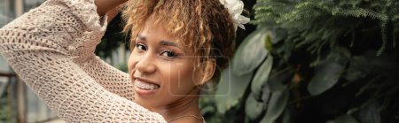 Modische und lächelnde afrikanisch-amerikanische Frau mit Hosenträgern, die im sommerlichen Stricktop posiert und in der Nähe grüner Pflanzen in der Orangerie in die Kamera blickt, Fashionista, die sich mit tropischer Flora vermischt, Banner 