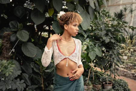 Stilvolle junge afrikanisch-amerikanische Frau im Sommer-Outfit schaut weg, während sie neben grünen Pflanzen im Hintergrund im Gartencenter steht, Fashionista mischt sich mit tropischer Flora