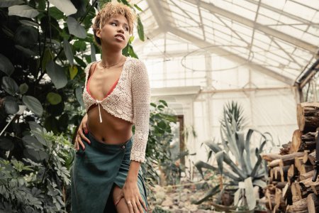 Mujer afroamericana joven de moda en traje de verano tocando la cadera y mirando hacia otro lado mientras está de pie en el centro del jardín borroso, mujer elegante con fondo tropical