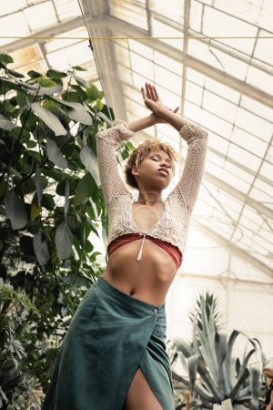 Low angle view of young african american woman in stylish knitted top and skirt posing with standing with closed eyes near green plants in orangery, stylish woman with tropical backdrop