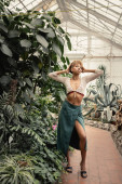 Full length of trendy young african american woman in summer knitted top and skirt standing and posing near green foliage in greenhouse, stylish woman with tropical backdrop t-shirt #663910272
