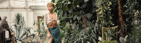 Young african american woman in knitted top and skirt posing and looking away while standing near green foliage in greenhouse, stylish woman with tropical backdrop, banner 