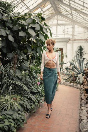 Pleine longueur de jeune femme afro-américaine en tenue d'été et talons marchant près de plantes vertes dans le centre de jardin, femme élégante avec toile de fond tropicale, concept d'été