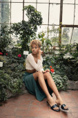 Full length of young african american woman in summer outfit and knitted top sitting near green plants with flowers in indoor garden, stylish woman with tropical backdrop, summer concept puzzle #663910386