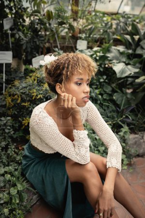 Portrait einer jungen und stylischen afrikanisch-amerikanischen Frau im Sommeroutfit, die sich in der Nähe grüner Pflanzen vor dem Hintergrund eines verschwommenen Gewächshauses entspannt, modebewusste Dame inmitten tropischen Grüns