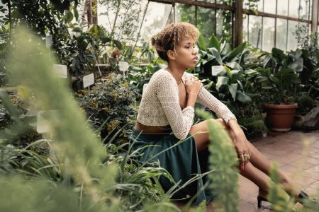 Mujer afroamericana joven y relajada en traje de verano y la parte superior de punto sentado cerca de plantas verdes en el jardín interior borroso en el fondo, señora de la moda hacia adelante en medio de la vegetación tropical