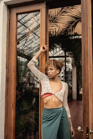Young african american woman in stylish summer outfit and knitted top standing near door of garden center and posing, fashionable woman enjoying summer vibes