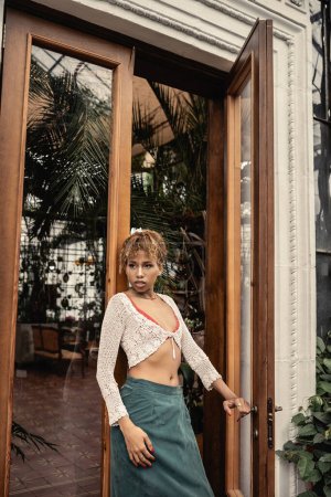 Young african american woman in summer outfit and knitted top opening door of indoor garden and looking away, fashionable woman enjoying summer vibes, botanical 