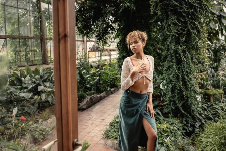 Joven y elegante mujer afroamericana en falda y verano de punto superior tocando el pecho mientras está de pie cerca de la puerta y las plantas en naranjería, señora con estilo rodeado de follaje tropical exótico 