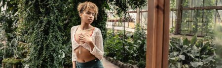 Élégante jeune femme afro-américaine en tricot haut et jupe regardant loin tout en se tenant près de la porte et des plantes dans le jardin intérieur, dame élégante entourée de feuillage tropical exotique, bannière 