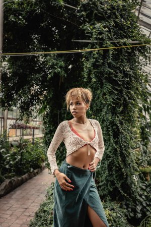 Selbstbewusste junge afrikanisch-amerikanische Frau im Sommeroutfit, die die Hand an der Hüfte hält und wegschaut, während sie neben Pflanzen in der Orangerie steht, stilvolle Dame umgeben von exotischem tropischen Laub