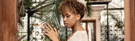 Young and stylish african american woman in knitted top touching brunch of palm tree and standing in blurred garden center at background, fashionista posing amidst tropical flora, banner 