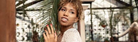 Positive young african american woman with braces wearing knitted top and standing near brunch of palm tree and looking away in blurred indoor garden, fashionista posing amidst tropical flora, banner