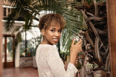 Portrait of stylish young african american woman with braces posing in knitted top touching brunch of palm tree and looking at camera in indoor garden, fashionista posing amidst tropical flora  magic mug #663911402