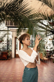 Young african american woman n stylish summer outfit touching branch of palm tree while standing in blurred indoor garden at background, fashionista posing amidst tropical flora  puzzle #663911454