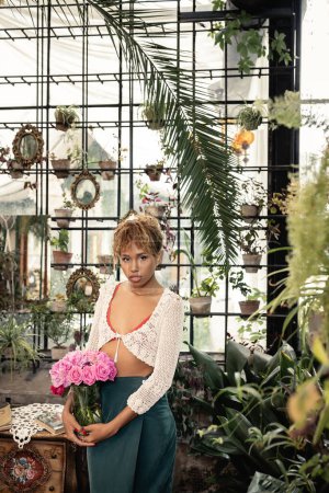 Mujer afroamericana joven de moda en traje de verano sosteniendo jarrón con rosas rosadas y mirando a la cámara cerca de las plantas en el jardín interior, mujer de moda con estilo tropical, concepto de verano