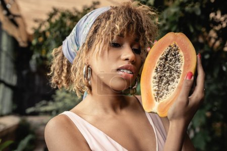 Porträt einer trendigen jungen afrikanisch-amerikanischen Frau in Kopftuch und sommerlichem Outfit mit Schnitt und reifer Papaya im verschwommenen Gartencenter, stilvolle Dame, die Mode und Natur vereint