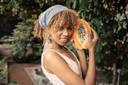 Zufriedene junge afrikanisch-amerikanische Frau mit Hosenträgern in Sommerkleid und Kopftuch, frische Papaya in der Hand und Blick in die Kamera in der Orangerie, stilvolle Dame, die Mode und Natur verbindet, Sommerkonzept