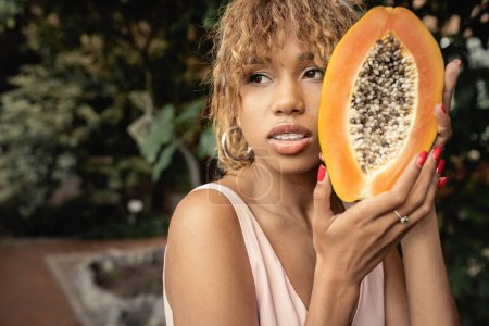 Confident young african american woman with braces and summer outfit holding fresh and ripe papaya and looking away in blurred greenhouse, stylish lady blending fashion and nature, summer concept