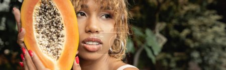 Portrait of trendy young african american woman with braces holding fresh and ripe papaya and looking away while spending time in indoor garden, stylish lady blending fashion and nature, banner 