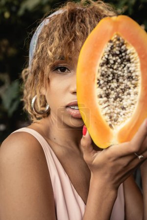 Retrato de mujer afroamericana joven y de moda en pañuelo para la cabeza y vestido de verano sosteniendo papaya cortada y madura y cubriendo la cara cerca de las plantas, elegante dama mezclando moda y naturaleza
